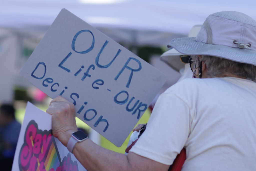 Person in white hat and shirt holds a hand-written sign "Our Life - Our Decision"